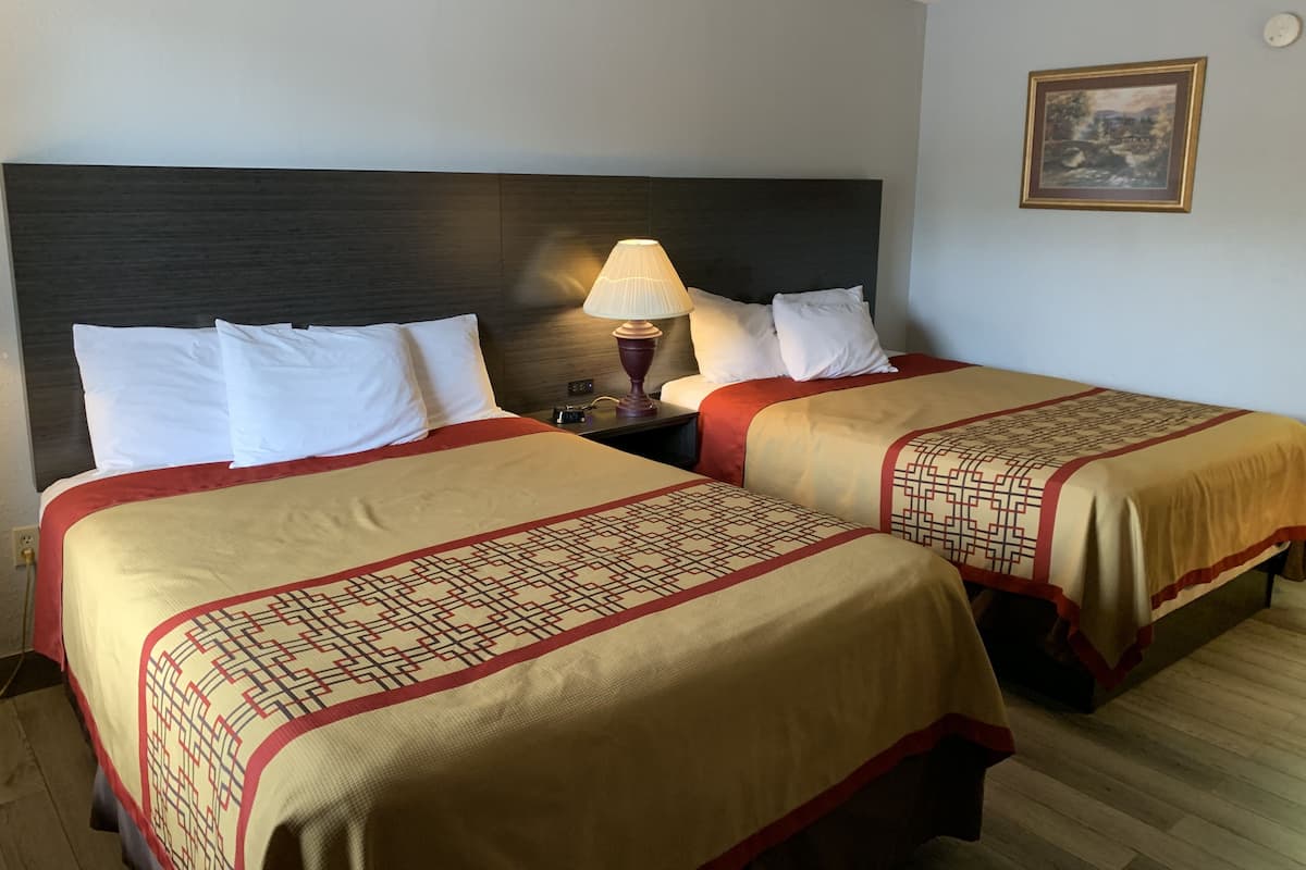 A hotel room with two beds, each with a red and beige bedspread, white pillows, and a nightstand with a lamp.