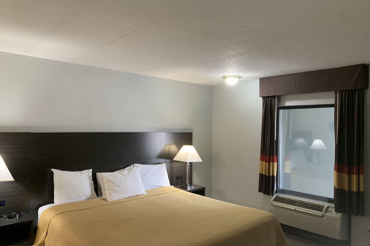 A hotel room with a bed, two bedside lamps, a window with curtains, and a wall-mounted air conditioner.