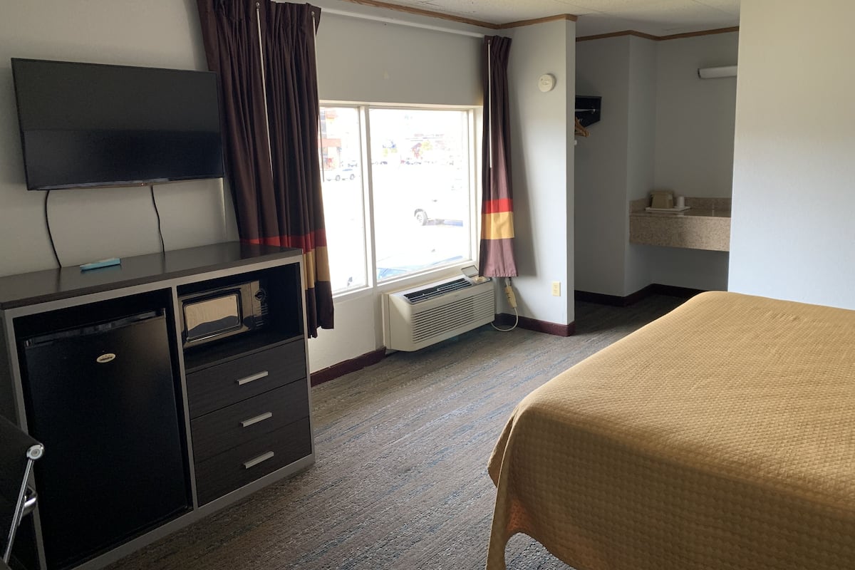 A hotel room with a bed, a TV on a dresser, a window with curtains, a radiator, and a small bathroom area.