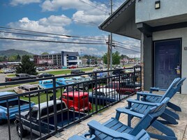 A balcony with blue chairs, a view of a parking lot with various vehicles, and a building with a dark door in the background.