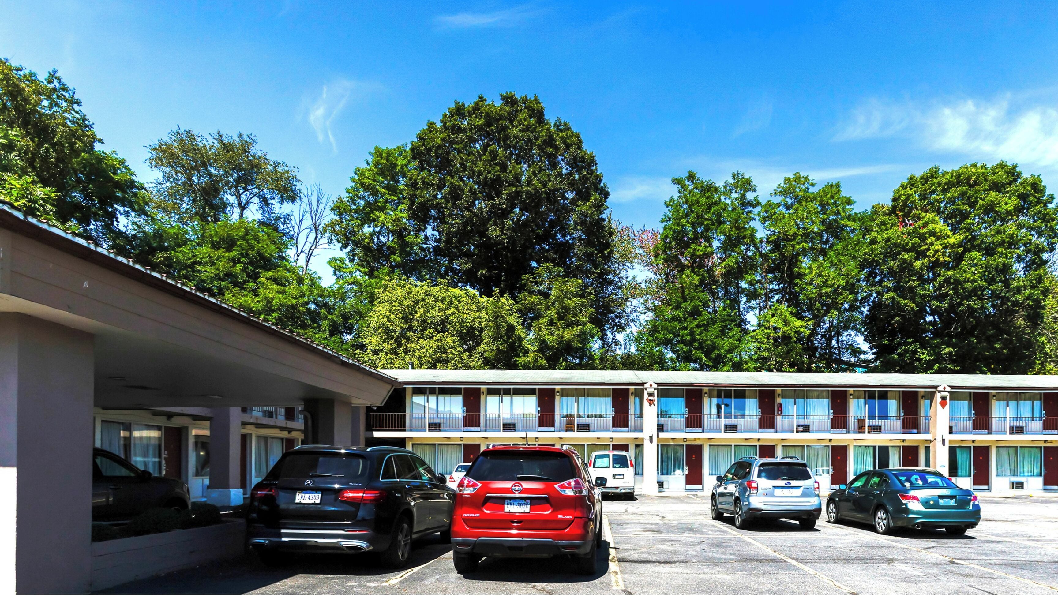 Un motel con una hilera de habitaciones, una zona de estacionamiento techada y árboles al fondo.