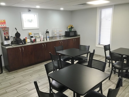 A modern kitchen with dark wooden cabinets, a granite countertop, and a microwave. There are two black tables with chairs, a window with blinds, and a vending machine.