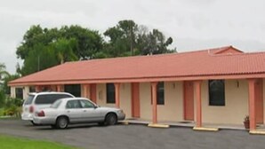 A single-story motel with a red roof and beige walls, featuring a parking area with two cars.