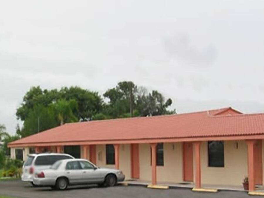 A single-story motel with a red roof and beige walls, featuring a parking area with two cars.