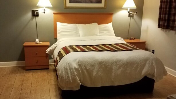 A neatly made bed with a white comforter and pillows, flanked by two bedside tables with lamps, a framed landscape painting above the bed, and a wooden floor.