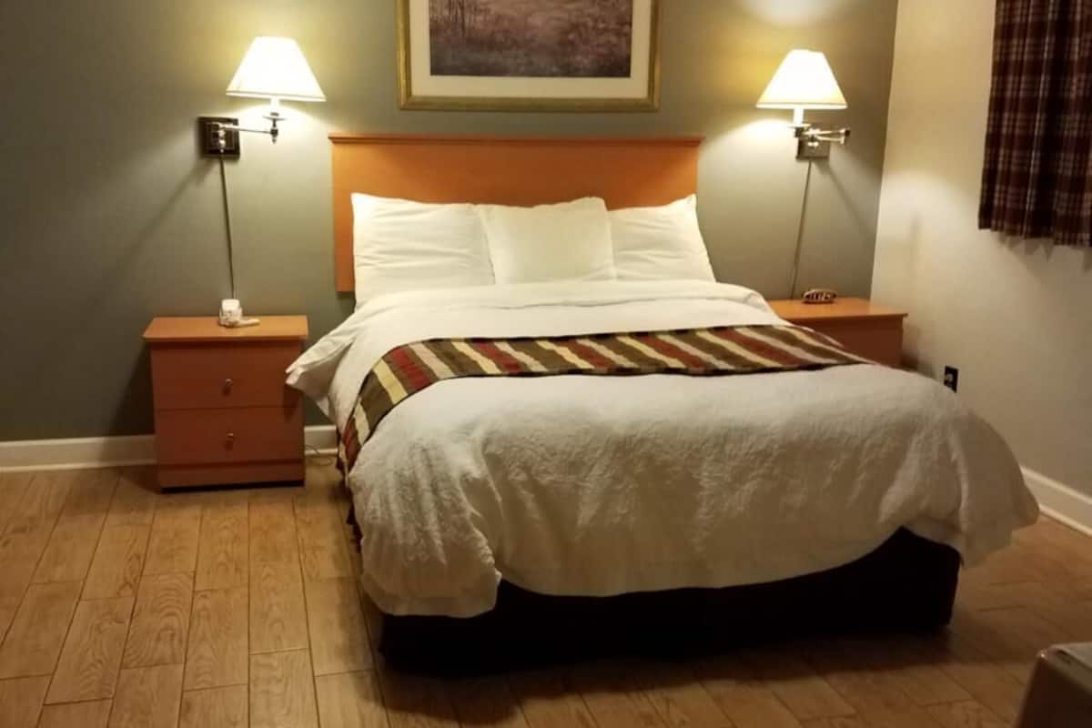 A neatly made bed with a white comforter and pillows, flanked by two bedside tables with lamps, a framed landscape painting above the bed, and a wooden floor.