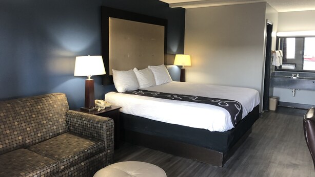 Ein Hotelzimmer mit einem großen Bett, zwei Nachttischlampen, einem gemusterten Sessel, einem kleinen Hocker und einem Badezimmer mit Waschbecken und Spiegel.