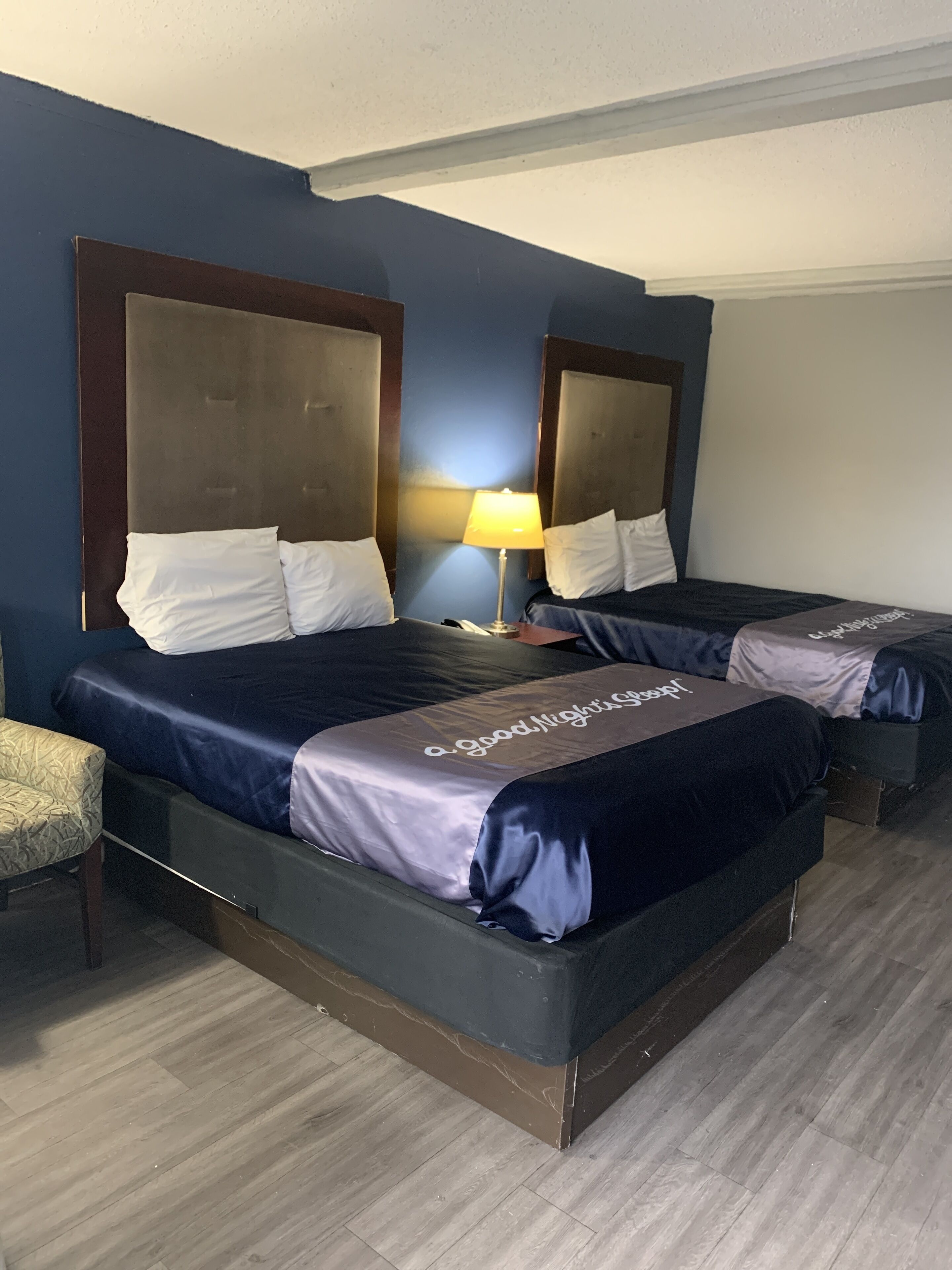 A hotel room with two beds, a nightstand with a lamp, and a chair.