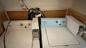 Deux machines à laver Whirlpool, placées l’une sur l’autre, dans une pièce dont le mur est recouvert de lambris en bois et où une prise électrique est visible.