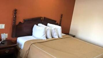 A hotel room with a wooden headboard, a bed with white linens and a beige blanket, a nightstand with a lamp, and a striped carpet.