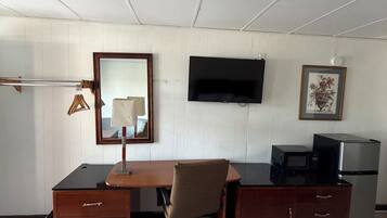 A room with a wooden desk, a chair, a television, a framed picture, a microwave, and a refrigerator.