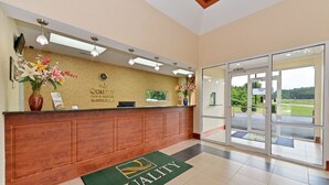 A hotel reception area with a wooden counter, a green 'Quality' mat, and a view of the outdoors through glass doors. - Quality Inn & Suites Cartersville North (White)
