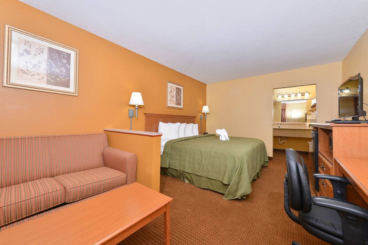 Une chambre d’hôtel avec un canapé, un lit recouvert d’une housse verte, un bureau avec une chaise, un miroir et des lampes fixées au mur.