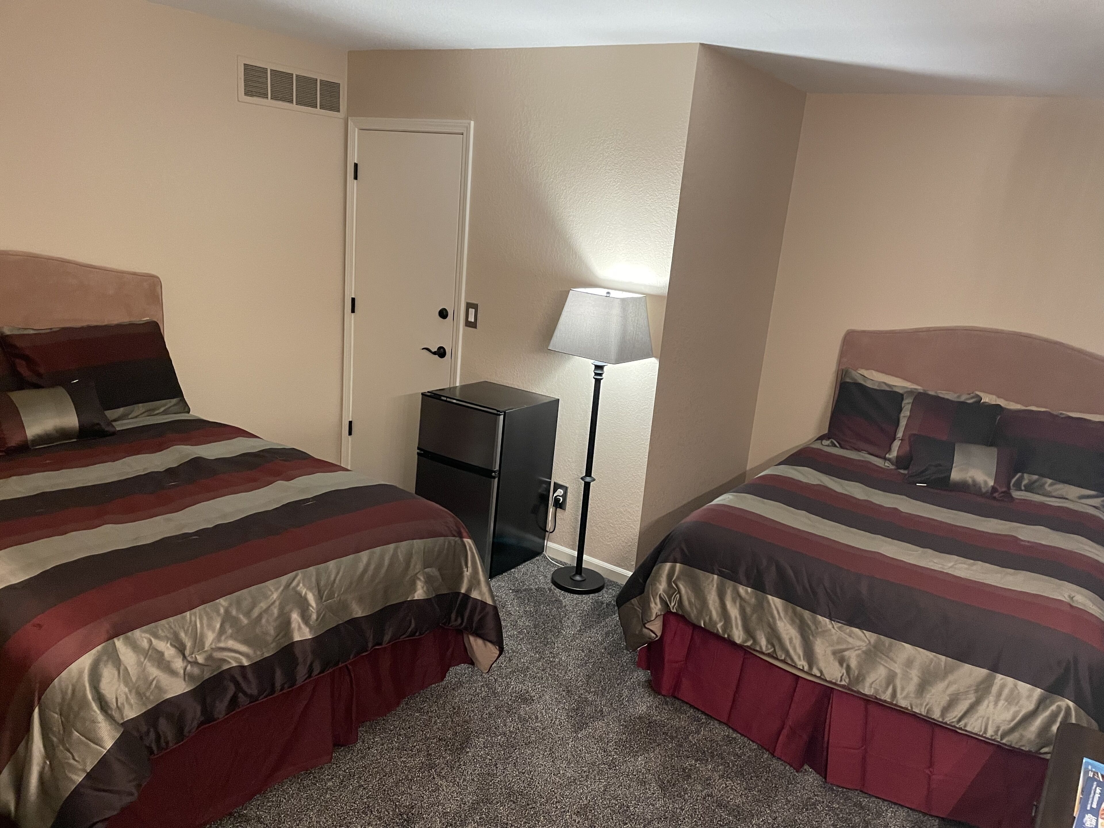 A hotel room with two beds, each with striped bedding, a small desk, a lamp, and a door.