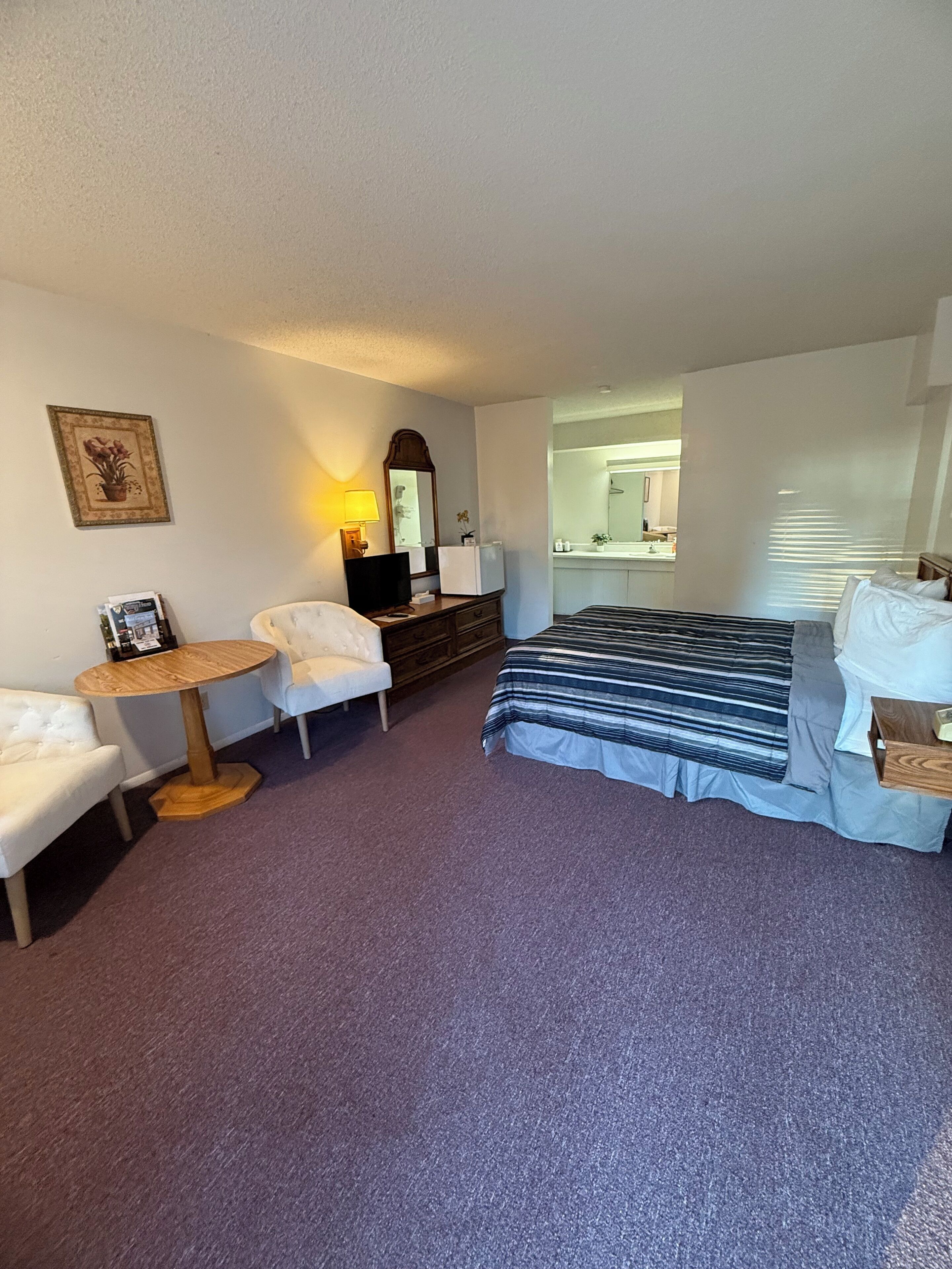 A hotel room with a bed, a desk, a chair, a small round table, a dresser, a mirror, and a bathroom visible through an open door.