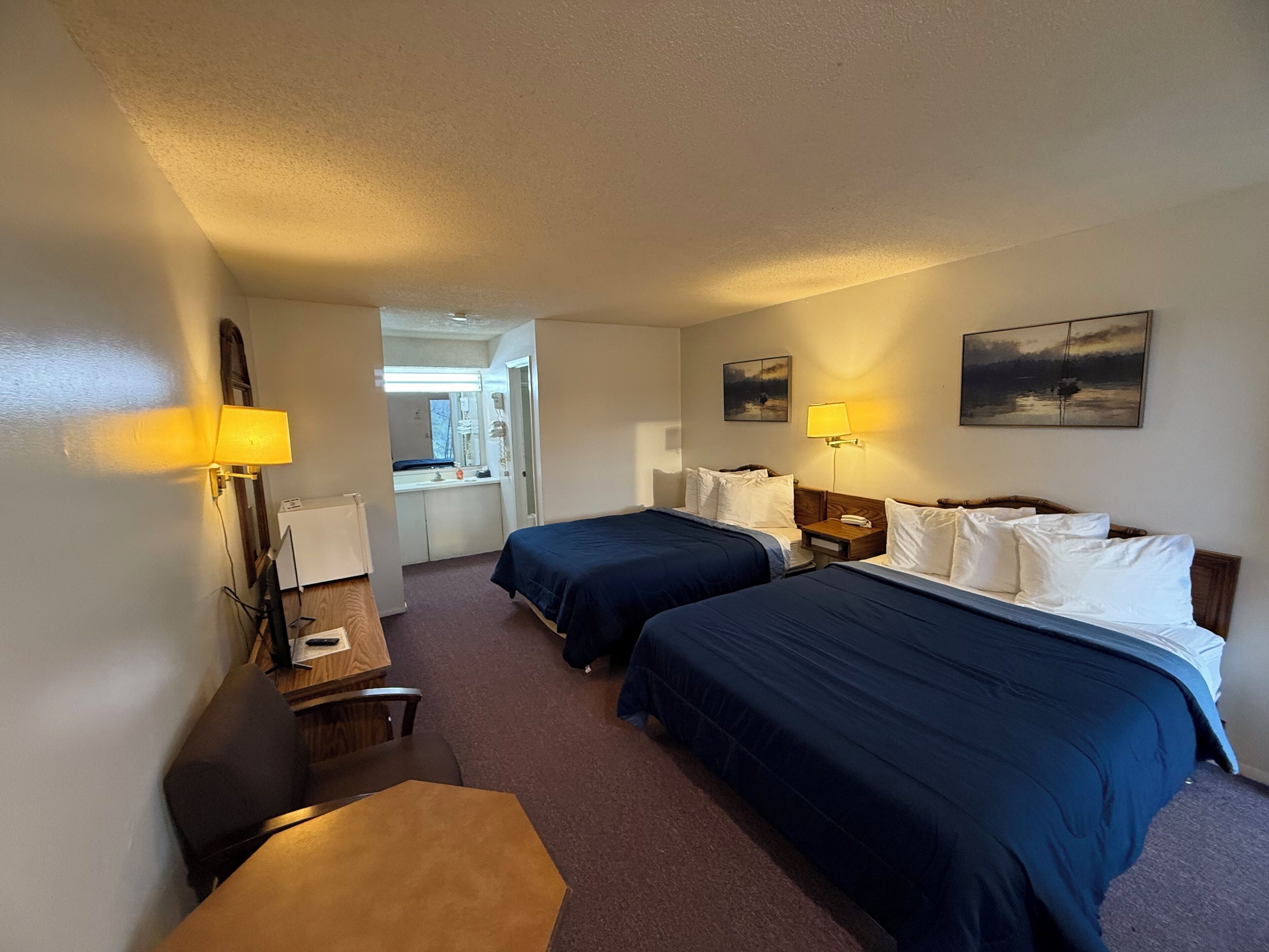 Une chambre d’hôtel avec deux lits, un bureau, une chaise et une salle de bain visible à travers une porte ouverte.
