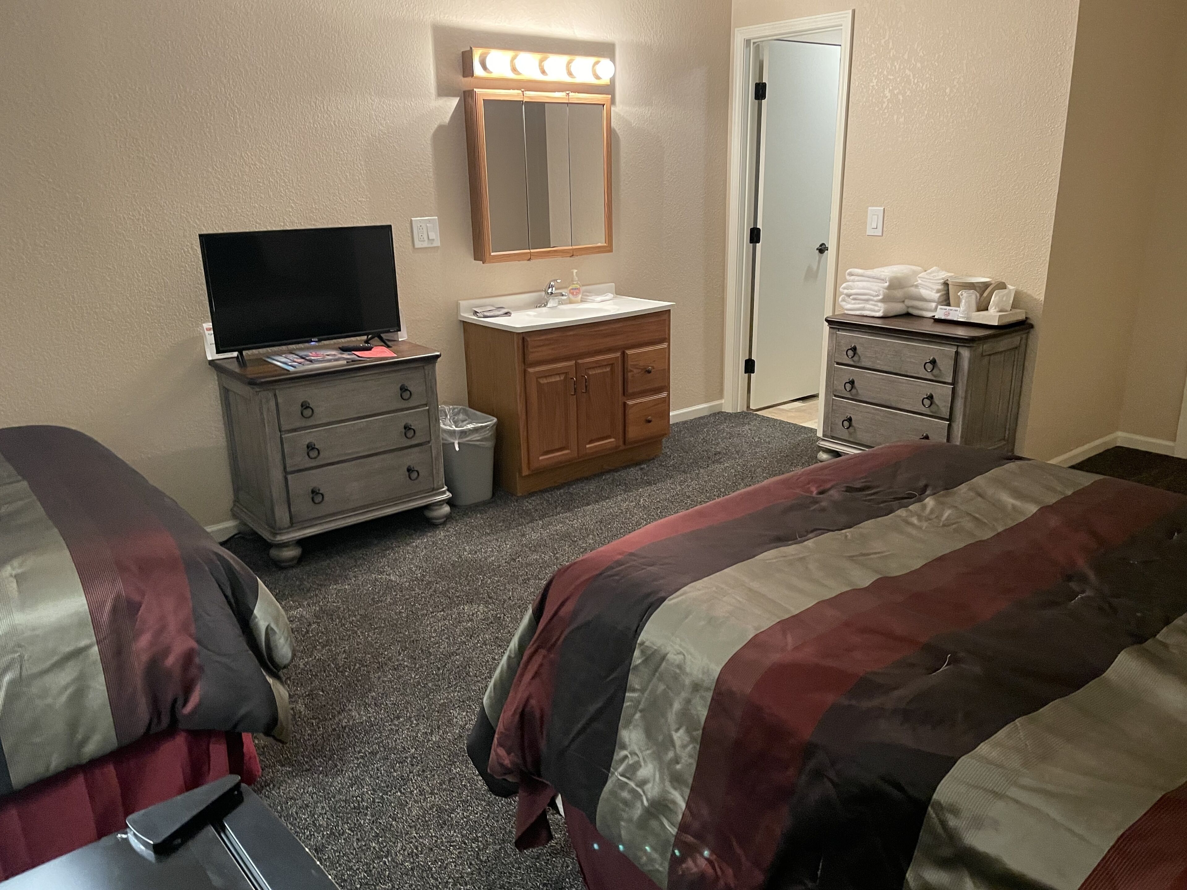 A hotel room with two beds, a vanity with a mirror, a television, and a door with a light above it.