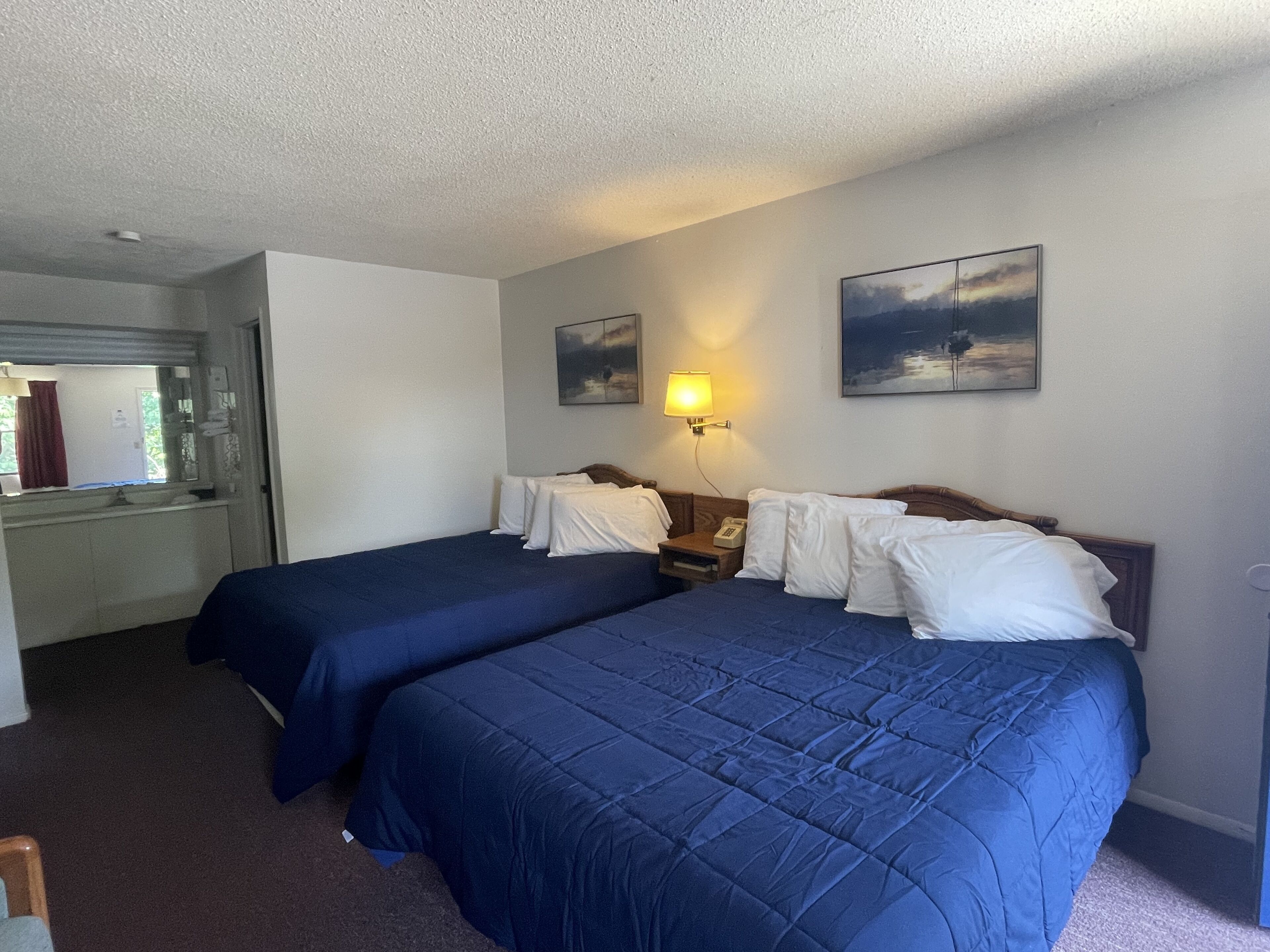 Une chambre d’hôtel avec deux lits, une table de chevet avec une lampe, un tableau au mur et une salle de bain visible à travers une porte ouverte.