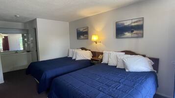 Une chambre d’hôtel avec deux lits, une table de chevet avec une lampe, un tableau au mur et une salle de bain visible à travers une porte ouverte.