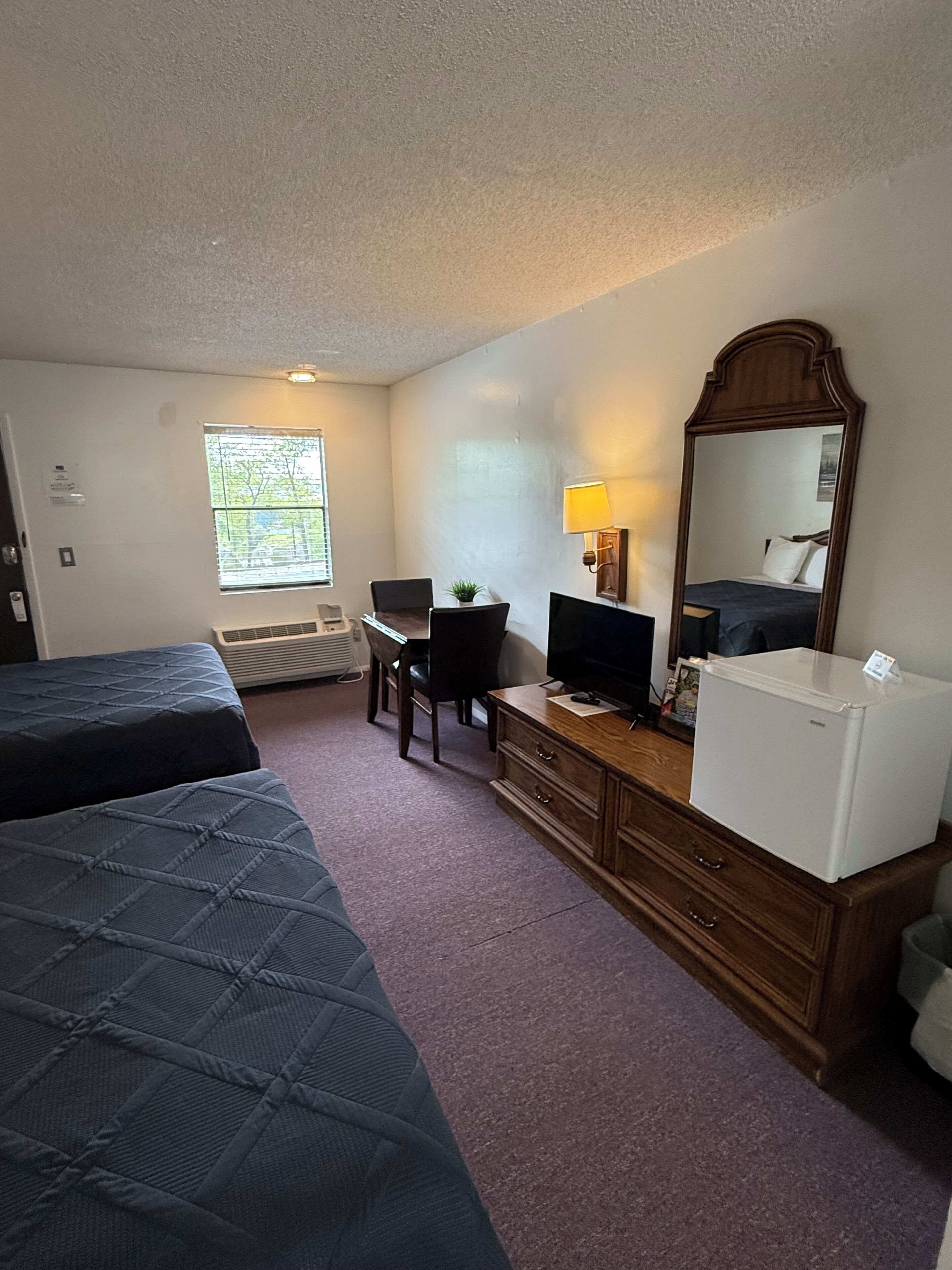 A hotel room with two beds, a desk with a computer, a refrigerator, a mirror, and a window with blinds.