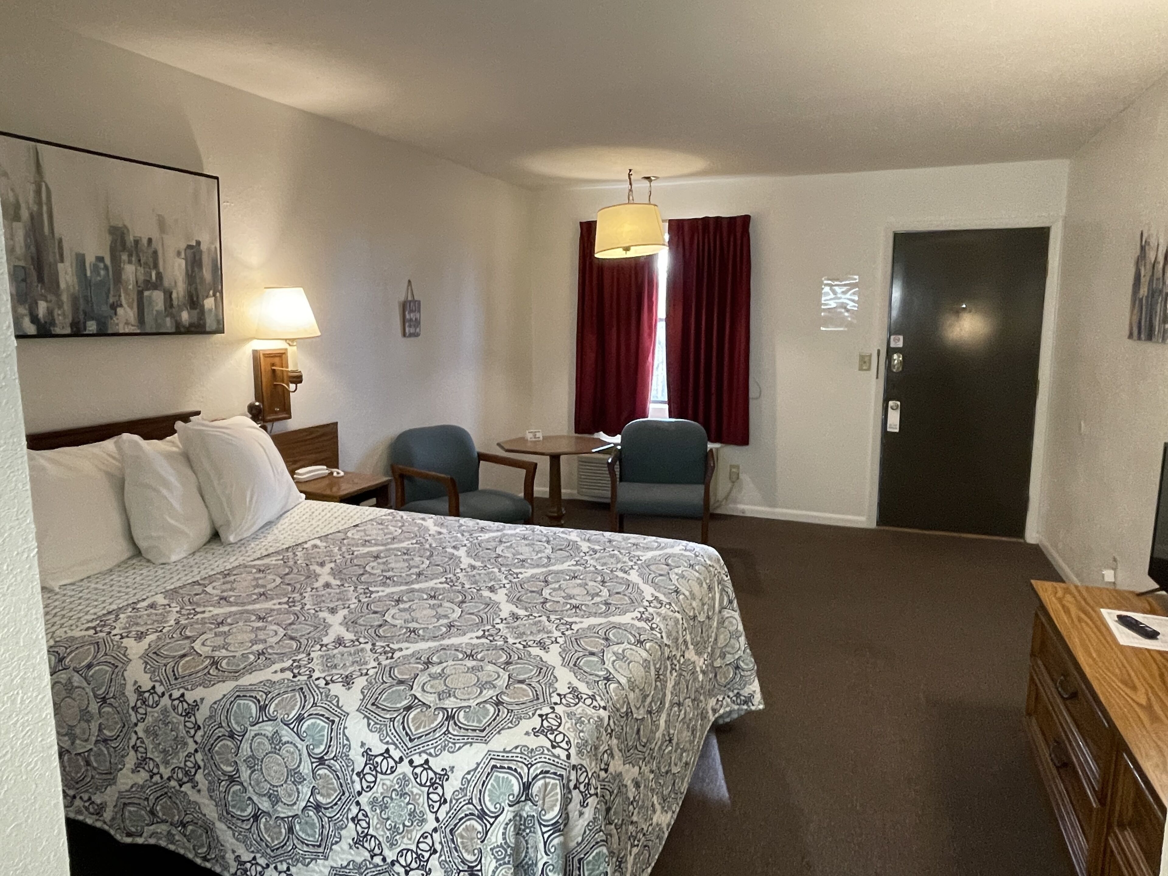 Ein Hotelzimmer mit einem großen Bett, einem Schreibtisch, zwei Stühlen, einer Lampe, einem Gemälde und einer Tür.