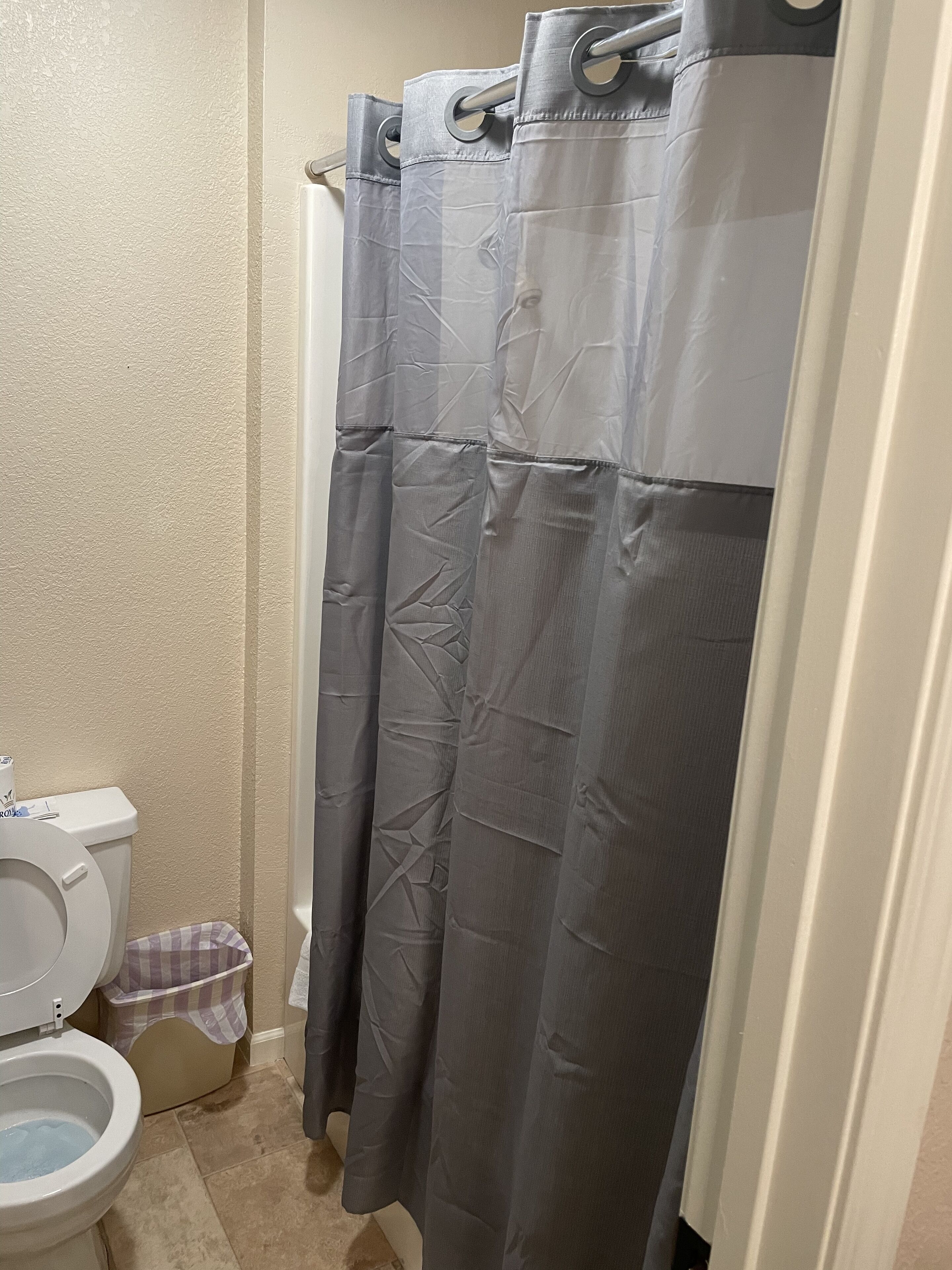 A bathroom with a toilet, a shower curtain, and a trash bin.