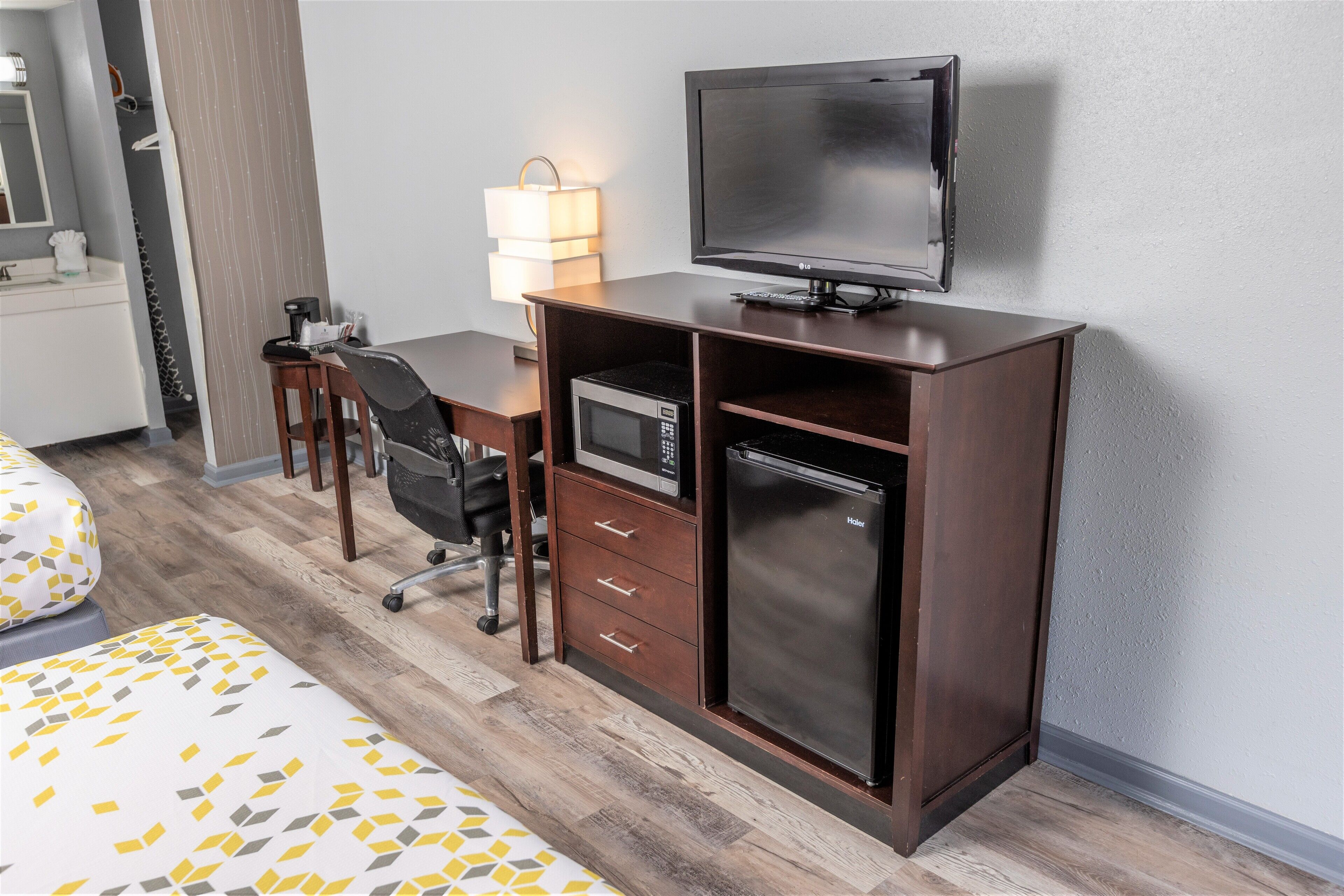 Habitación de hotel con un escritorio de madera, un televisor de pantalla plana, microondas, refrigerador pequeño, una silla, una lámpara y una cama con colcha estampada.