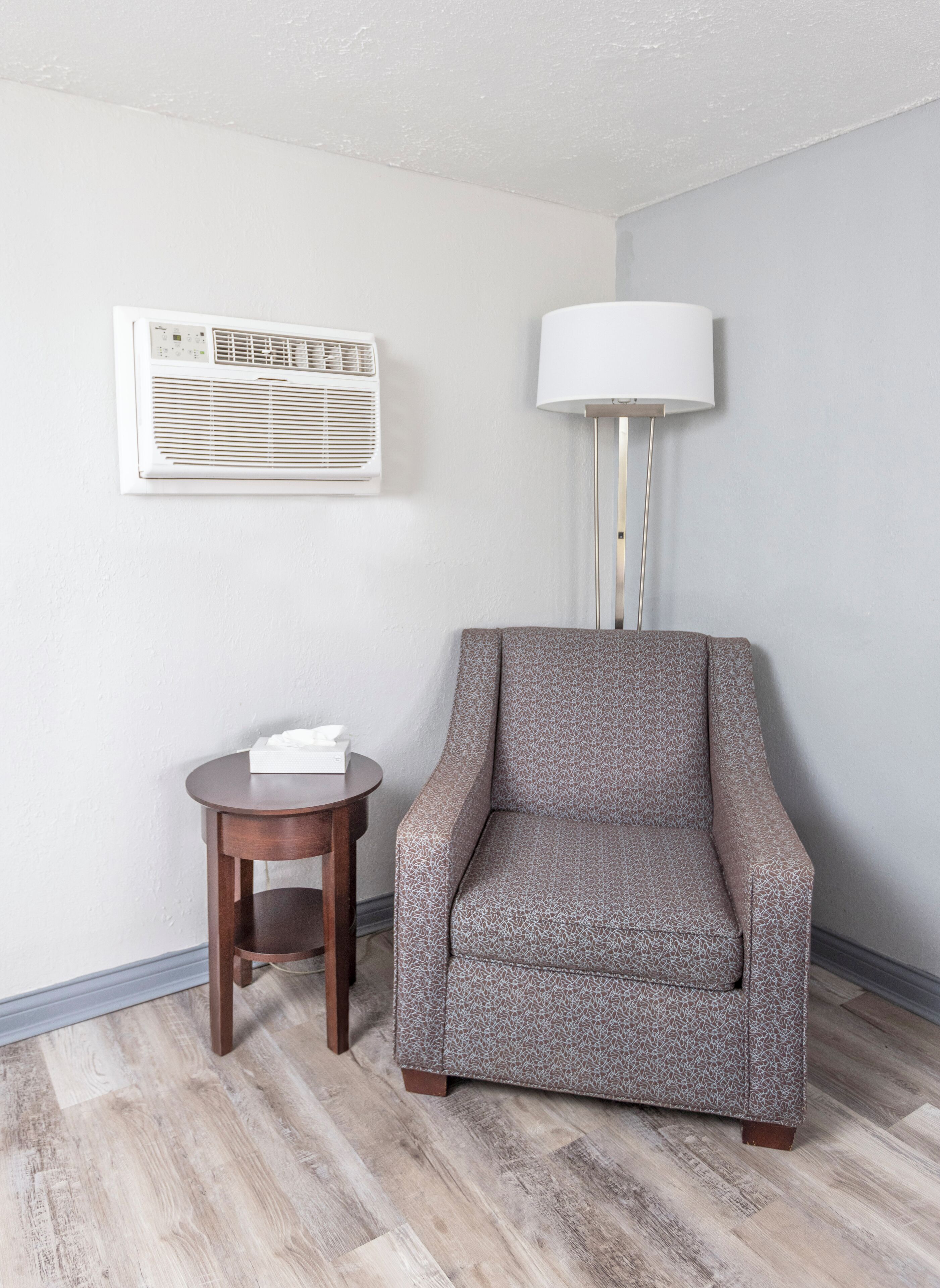 Una habitación con un sillón estampado, una mesita de madera, una lámpara blanca y un aire acondicionado de pared.