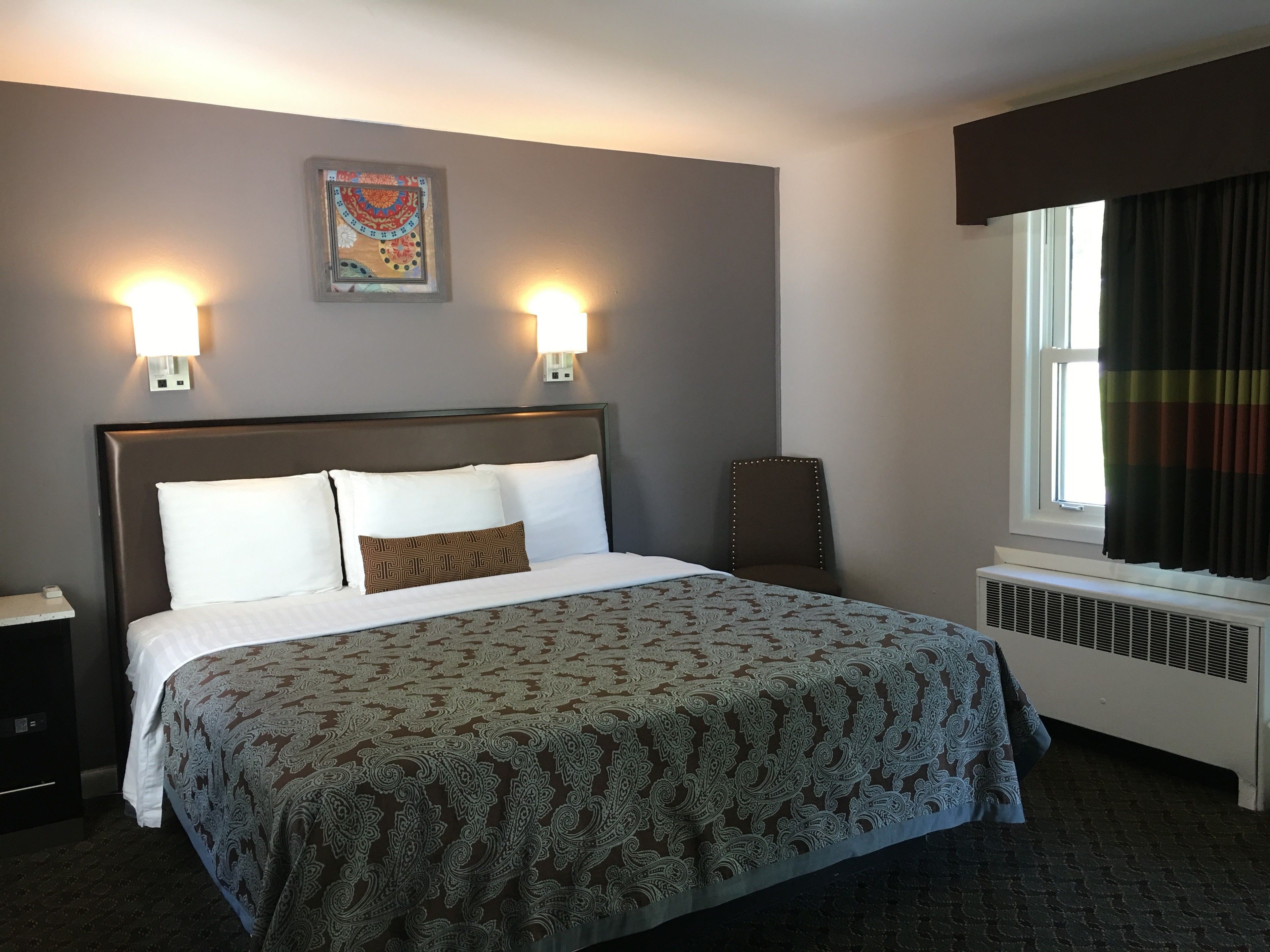 A neatly made bed with a patterned bedspread, two wall-mounted lights, a decorative wall hanging, and a window with curtains.