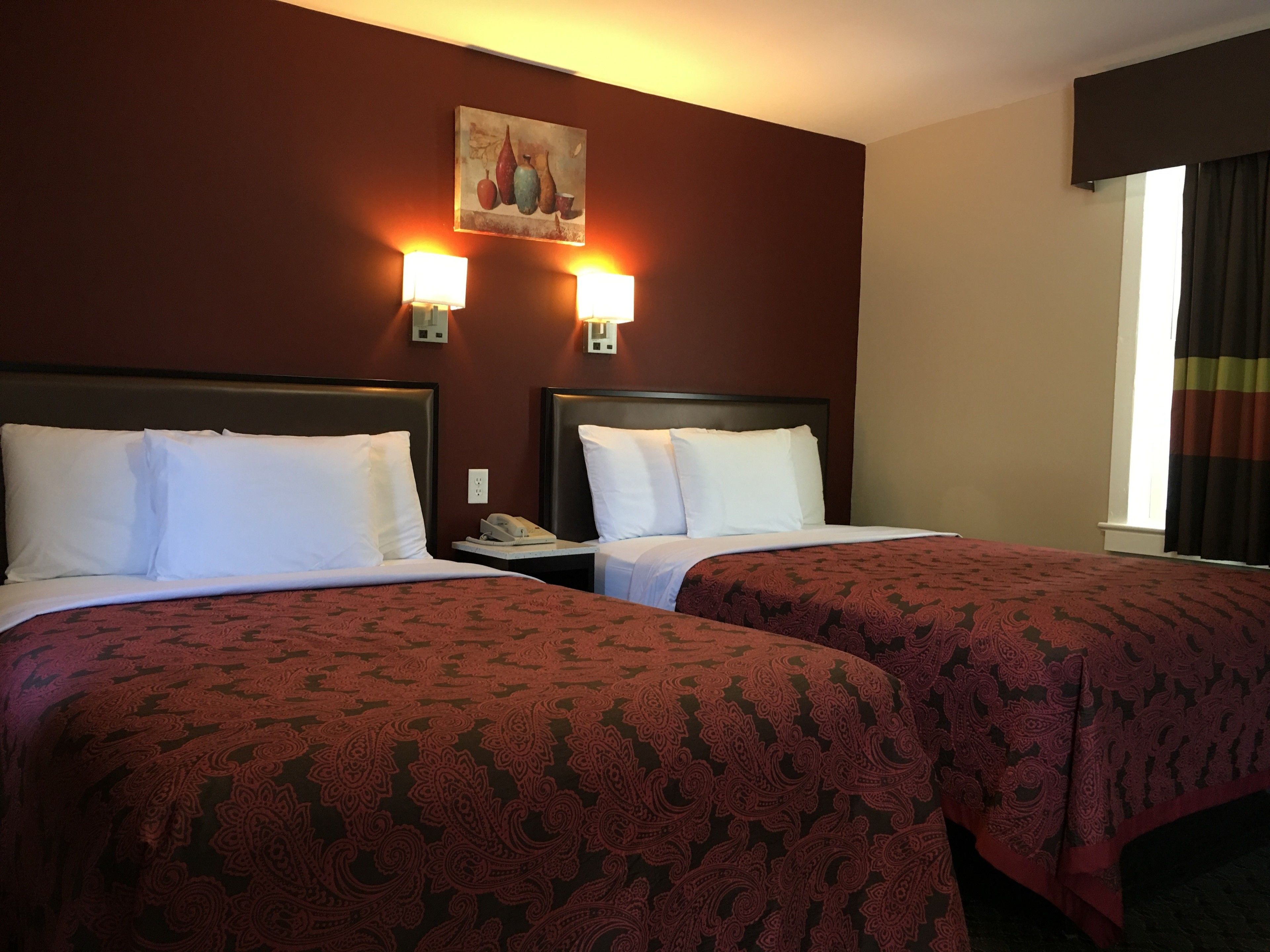 Une chambre d’hôtel avec deux lits, chacun recouvert de draps blancs et d’une couvre-lit à motifs rouges. De chaque côté d’un tableau placé au-dessus des lits, on trouve des lampes fixées au mur.