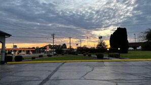 Un estacionamiento con una gasolinera al fondo, rodeado de vegetación y un cielo nublado.