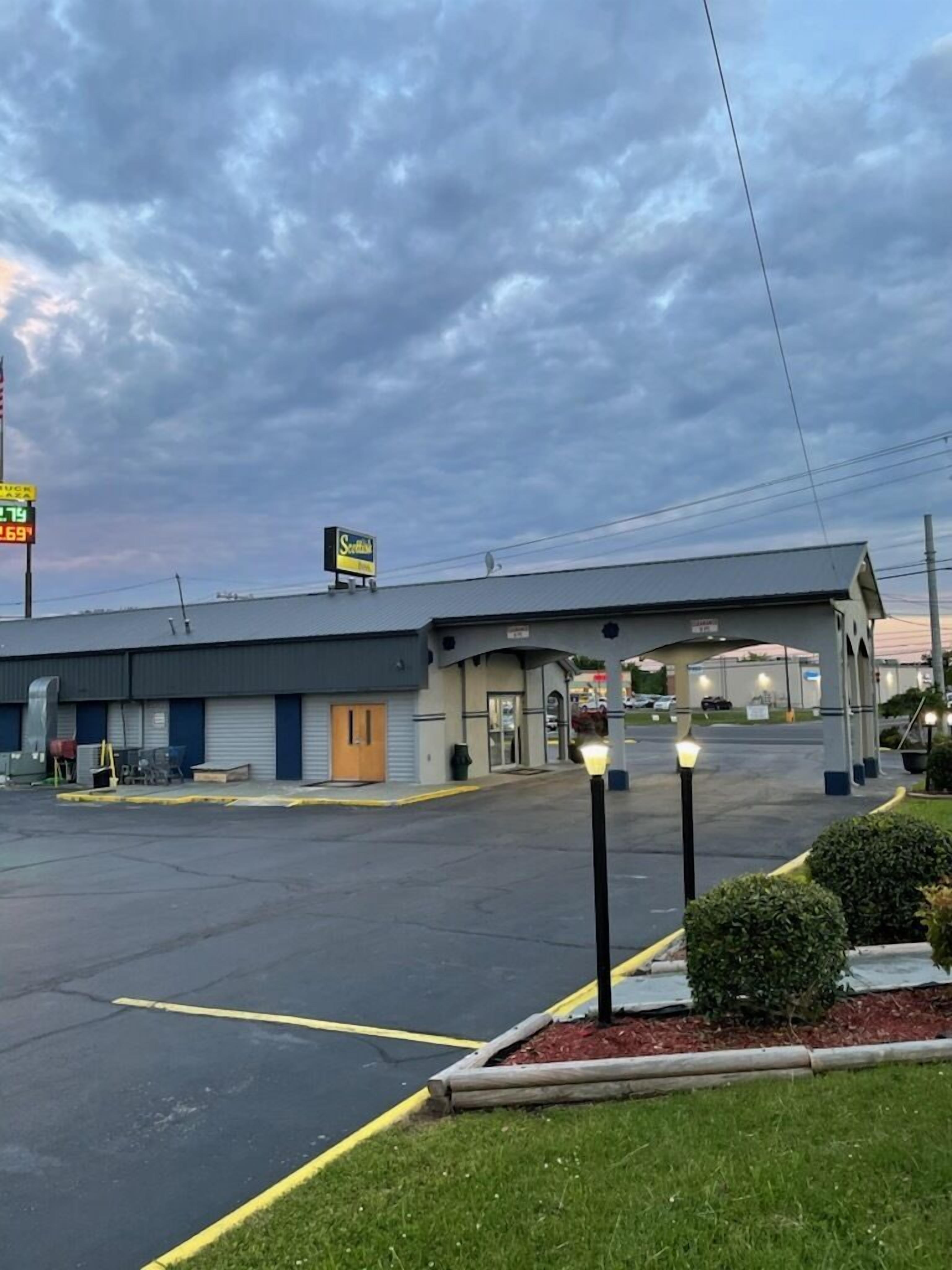 Ein Motel mit eingeschossigem Bau, überdachtem Parkplatz und einem kleinen begrünten Bereich mit einer Laterne.