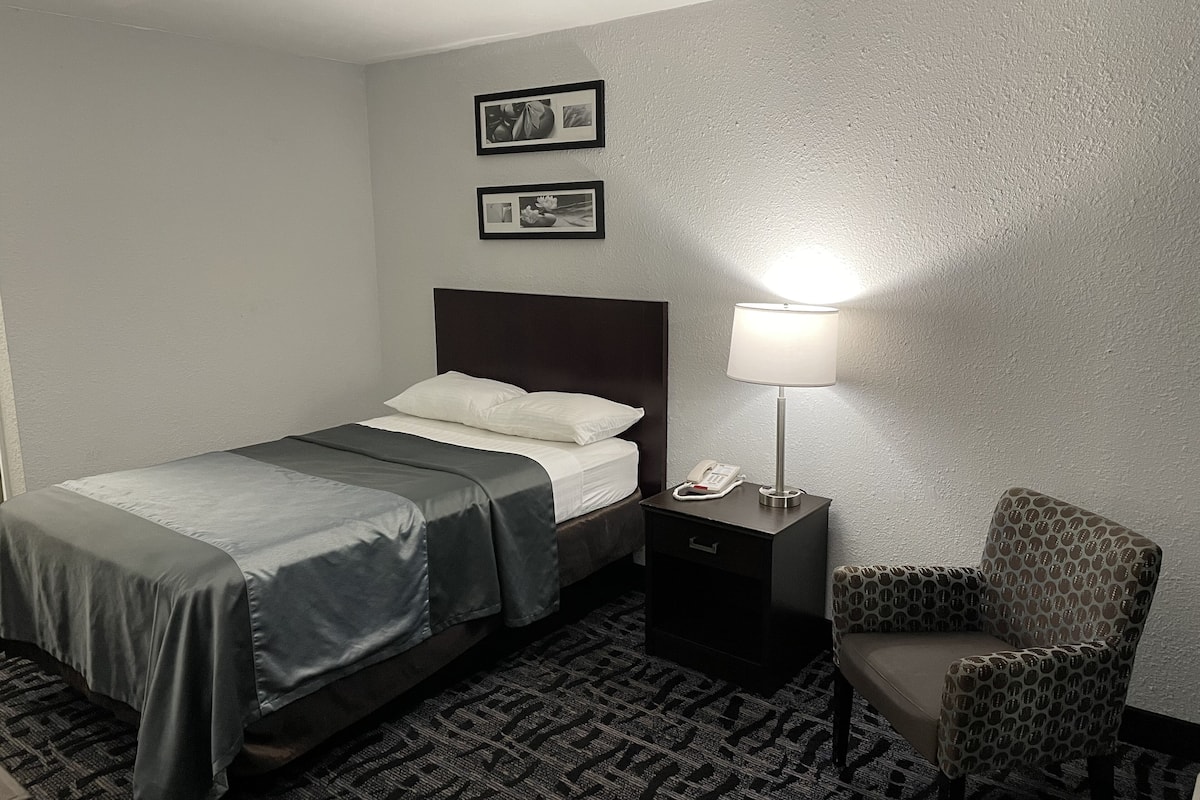 A hotel room with a bed, a bedside table with a lamp, a chair, and two framed pictures on the wall.