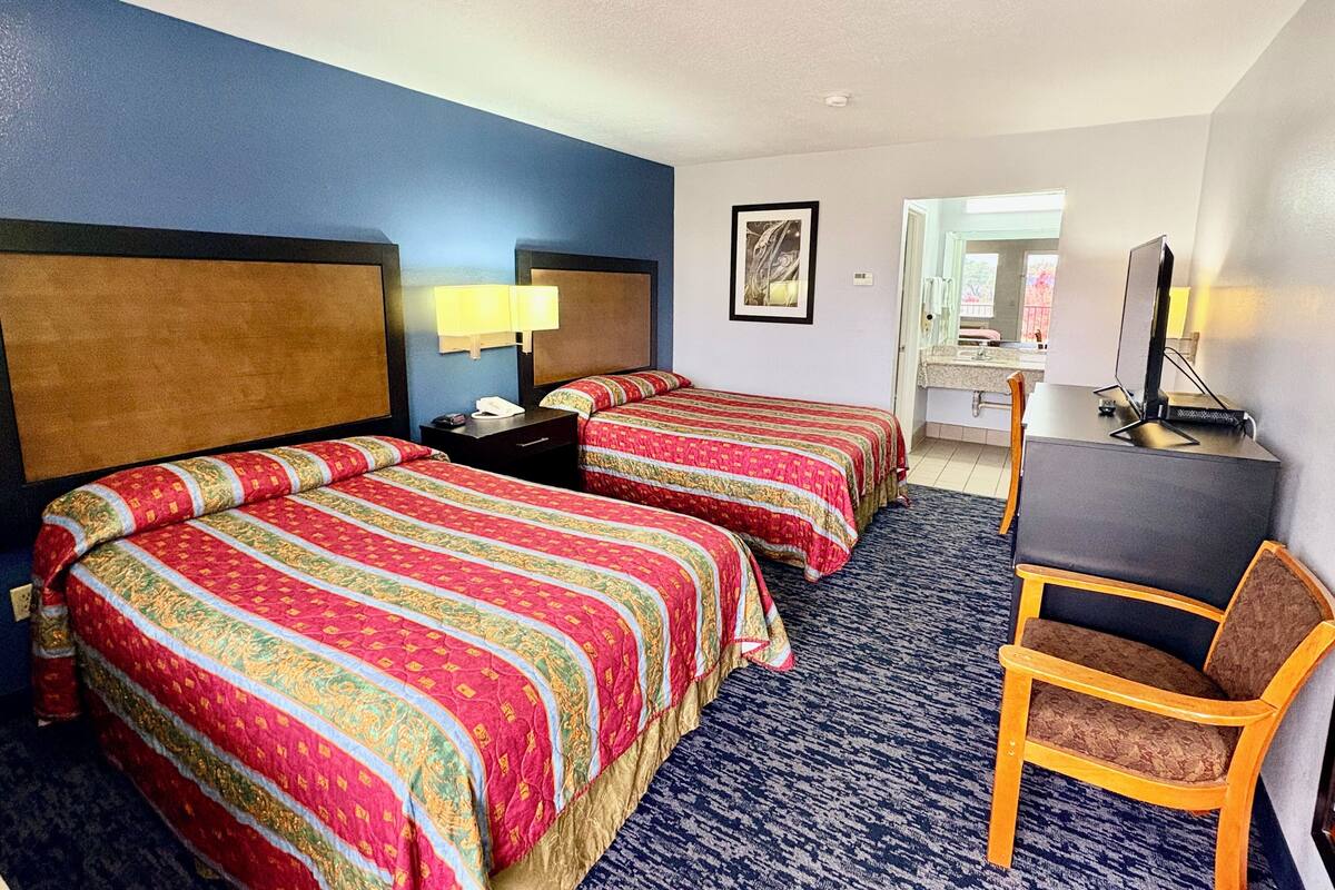 A hotel room with two beds, a desk, a chair, a lamp, and a bathroom visible through an open door.