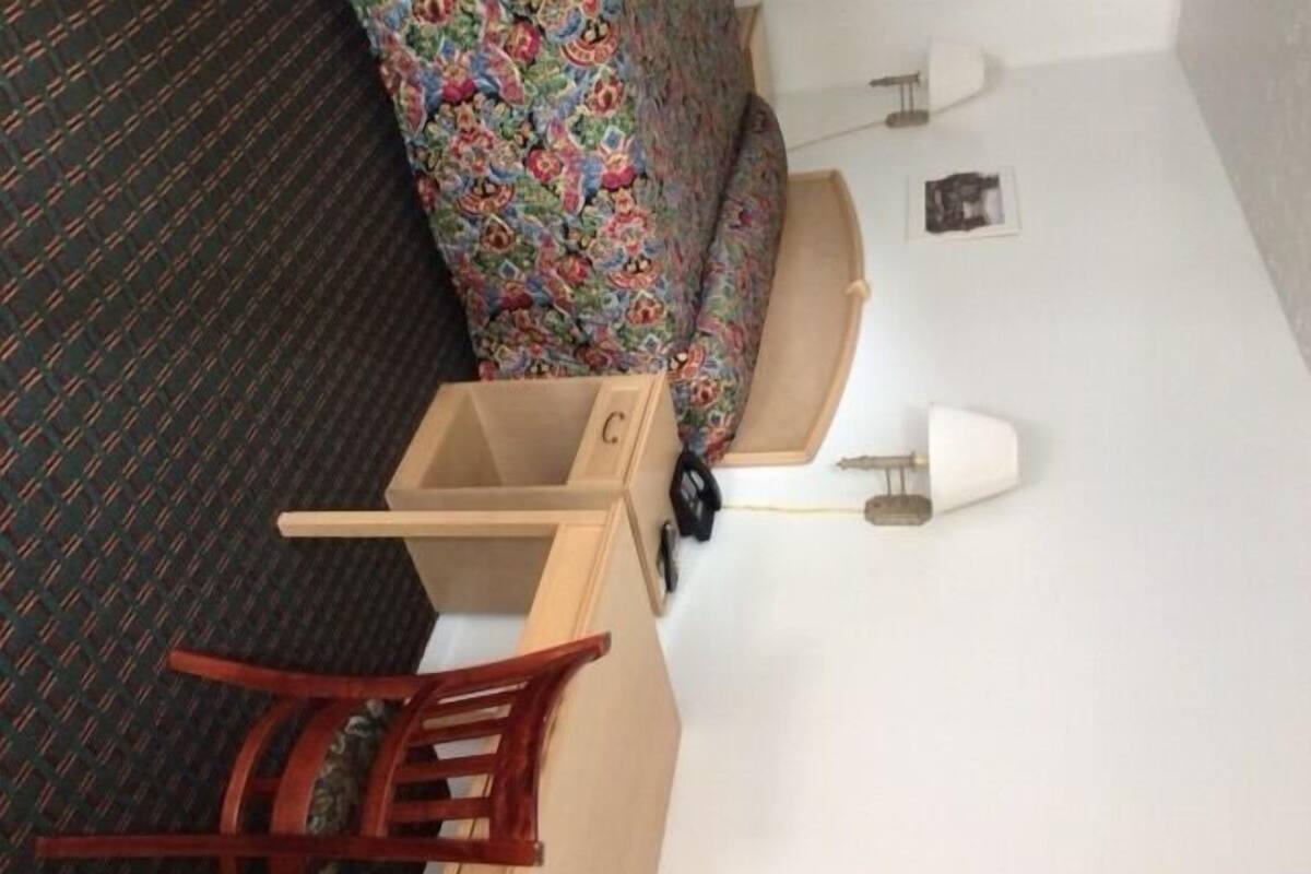 A small wooden chest with a drawer, a wooden chair, a colorful patterned sofa, and a white lamp on a white floor.