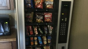 A vending machine stocked with snacks and beverages, located in a kitchen area with wooden cabinets and a microwave.