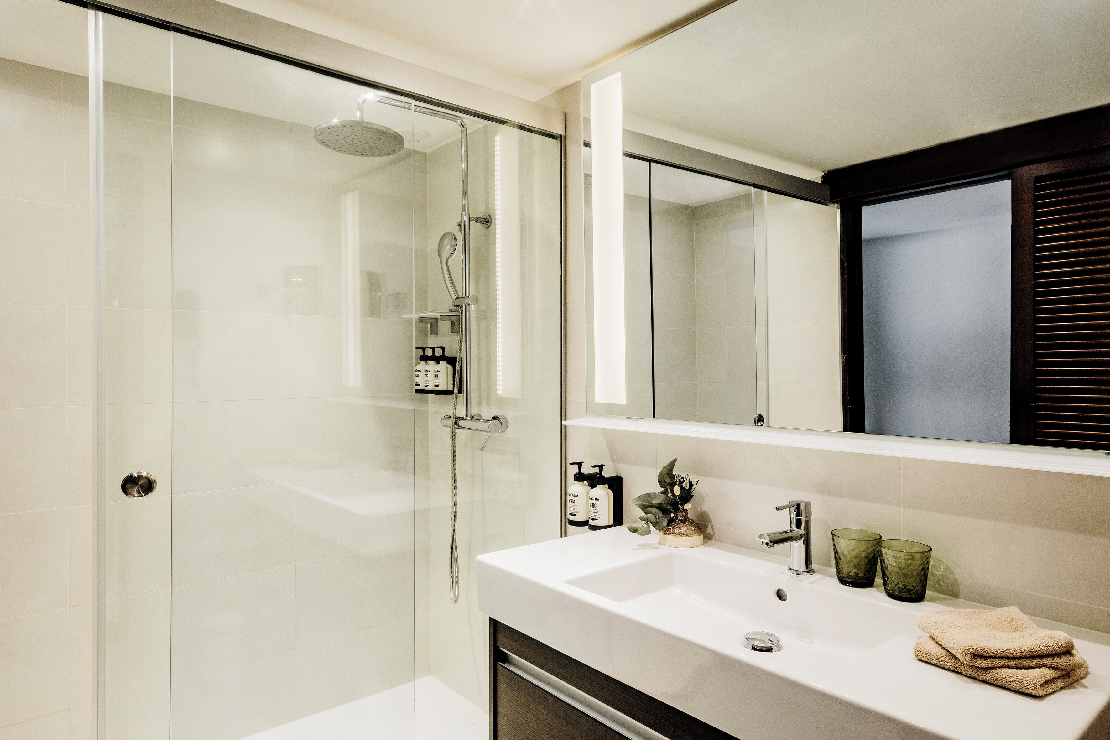 A modern bathroom with a glass-enclosed shower, a white sink with two green glasses, and a towel on the counter.