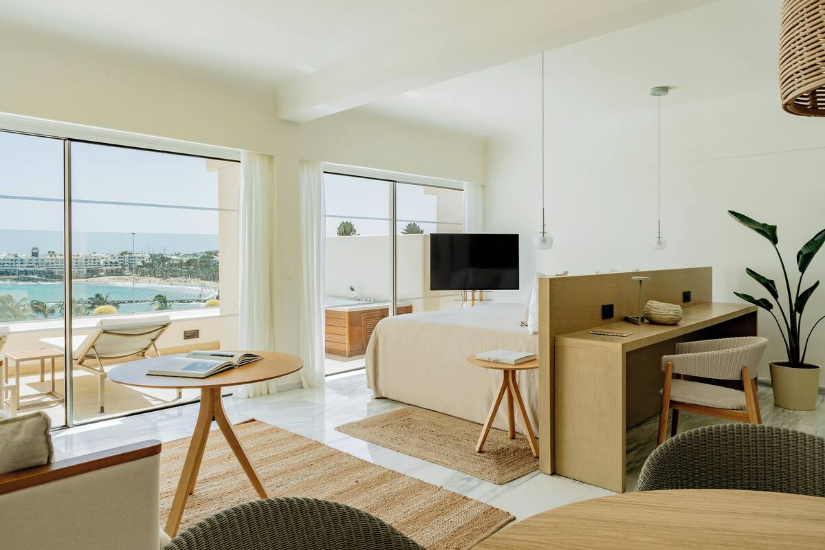 A modern hotel room with a large bed, a desk, a TV, and a balcony with a view of the ocean.