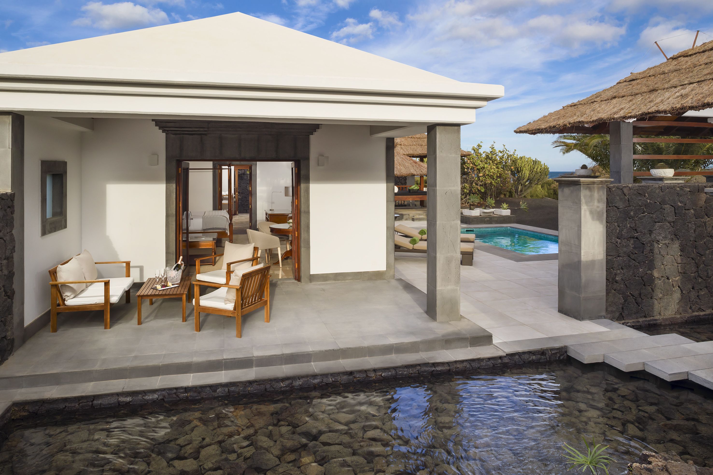 Una villa moderna con piscina, mobiliario exterior y una estructura con techo de paja.