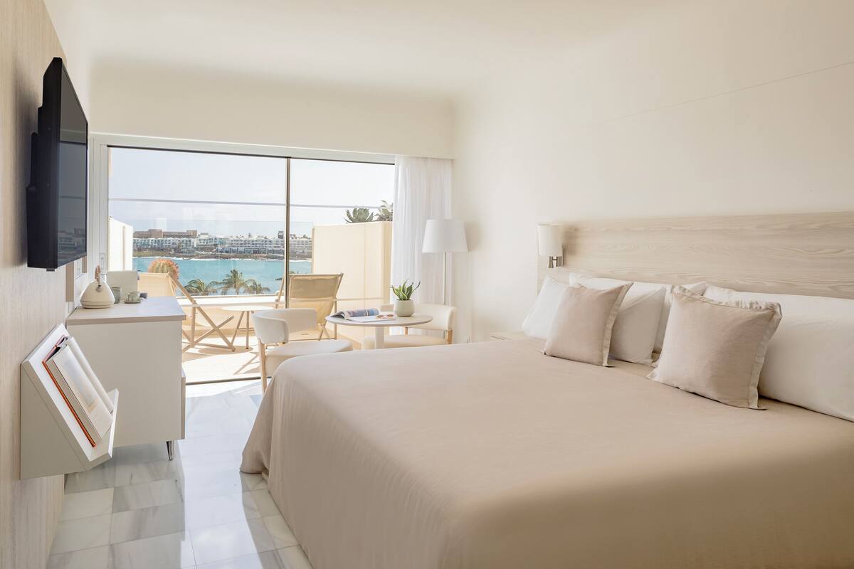 A hotel room with a large bed, a desk, a chair, a small table, and a view of the ocean.