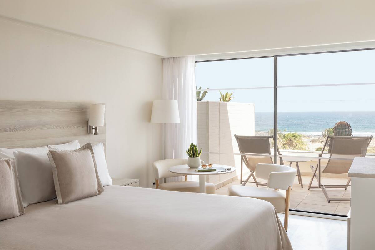 A hotel room with a large bed, a small table with a plant, and a view of the ocean through large windows.