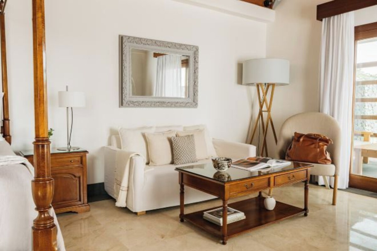 A hotel room with a wooden bed, a white sofa, a glass coffee table, a mirror, and a lamp.