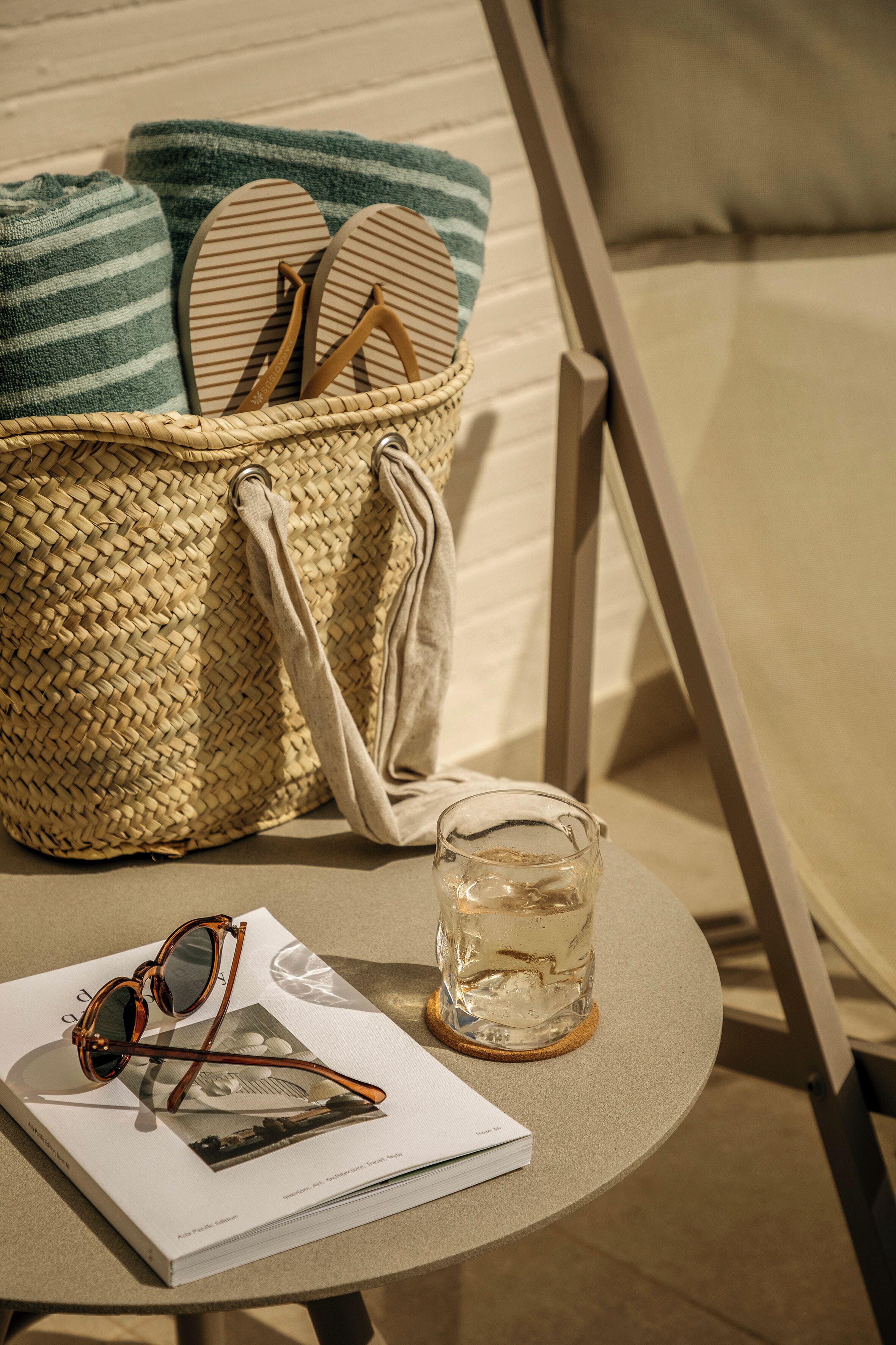 A wicker basket with flip-flops and a towel, a glass of water, sunglasses, and a magazine on a round table.