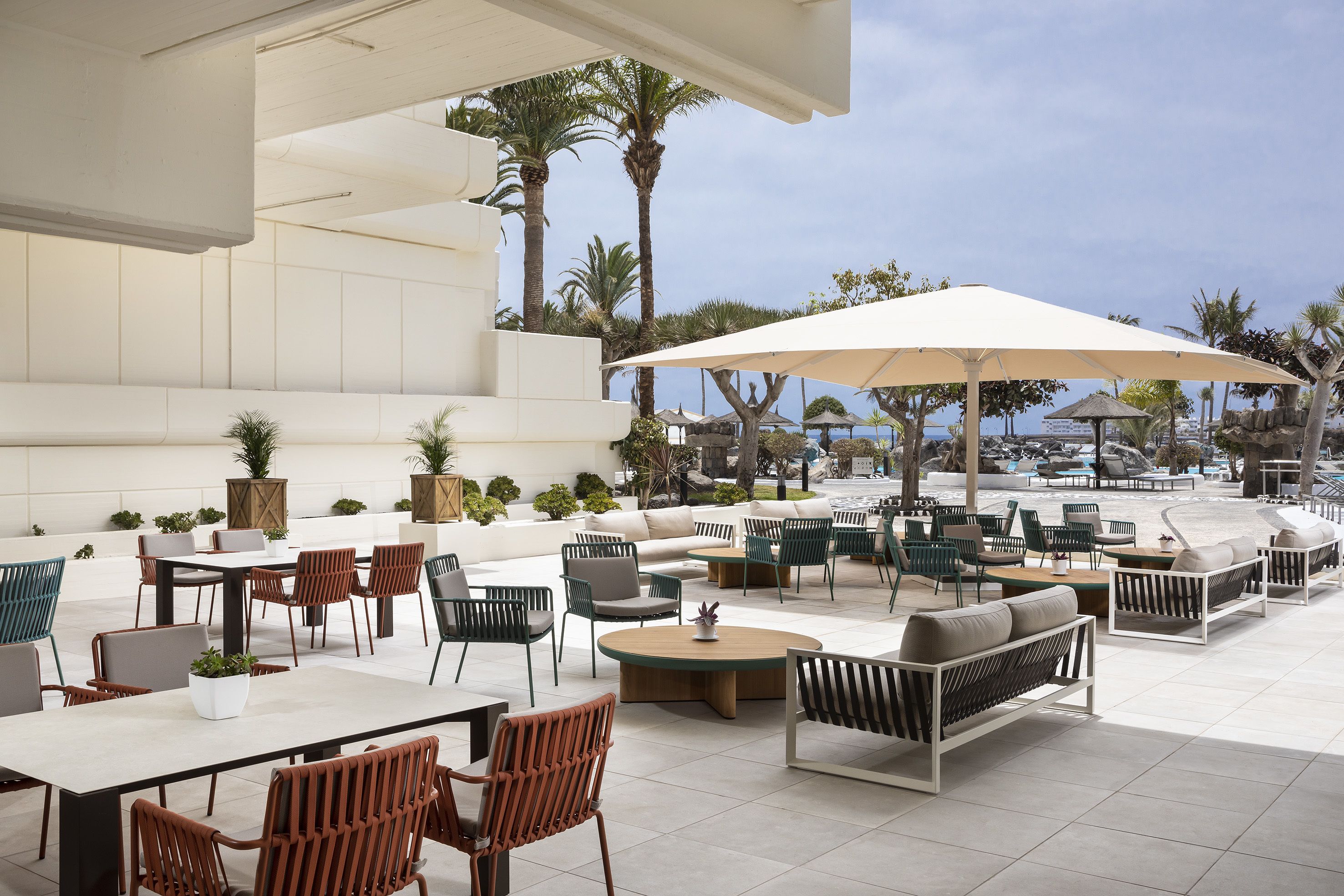 Een terras buiten met zithoekjes, tafels en stoelen, omringd door potplanten en parasols.