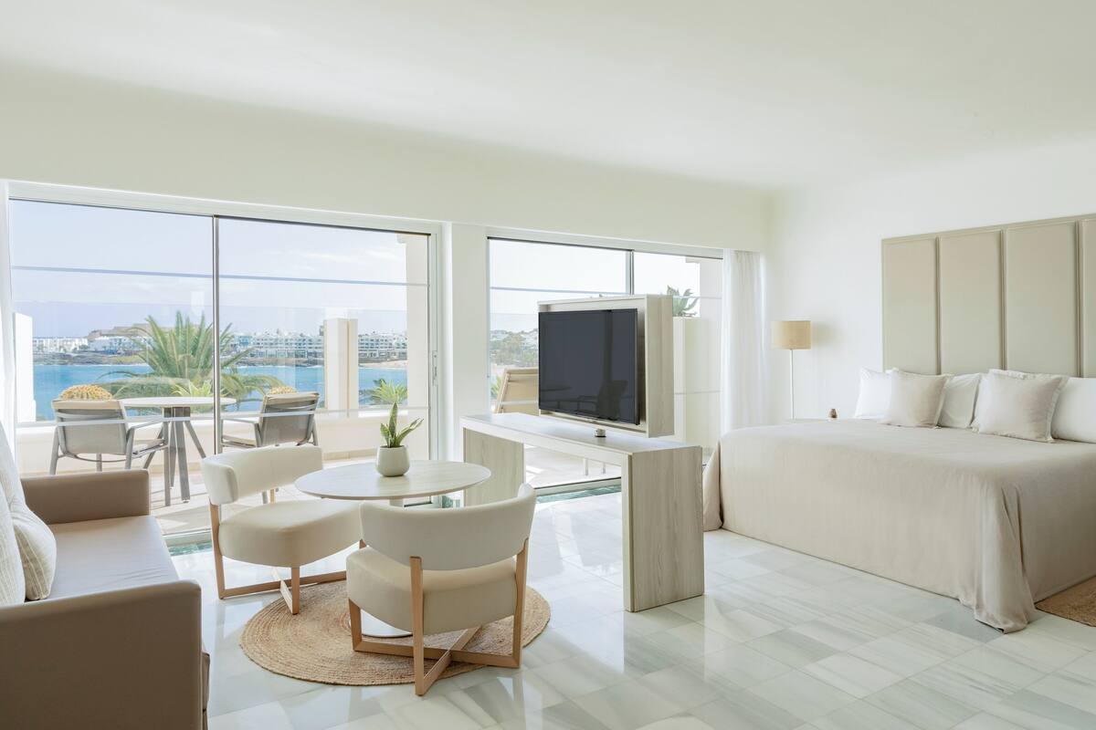 A modern hotel room with a large bed, a TV, a dining area with a table and chairs, and a balcony with a view of the city.