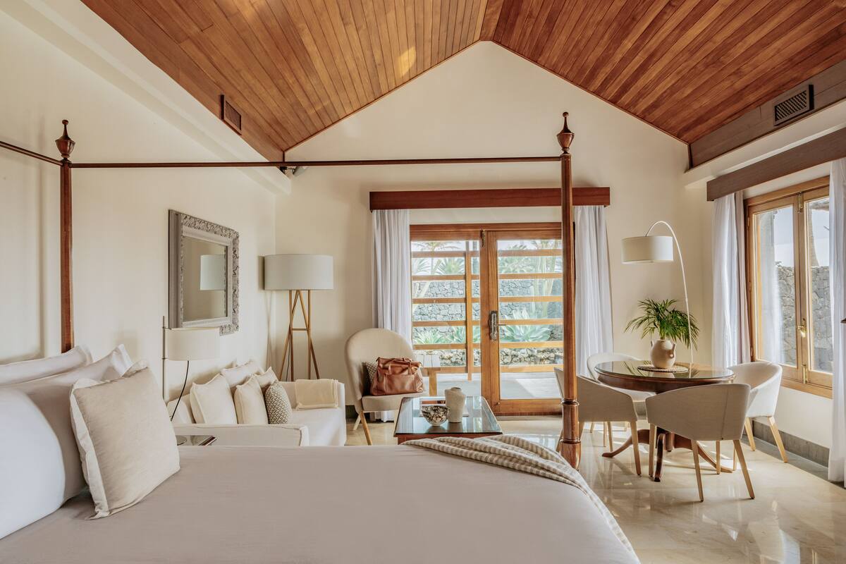 A spacious bedroom with a large bed, a sitting area with a table and chairs, and a view through a glass door to the outside.