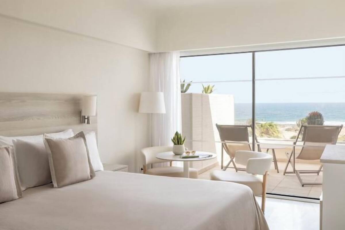 A hotel room with a bed, a small table with chairs, a lamp, and a view of the ocean.