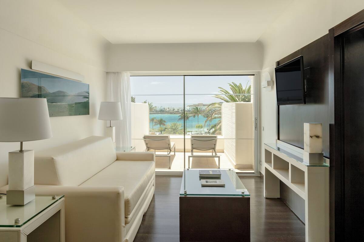 A modern hotel room with a sofa, two chairs, a coffee table, a TV, and a balcony with a view of the sea.