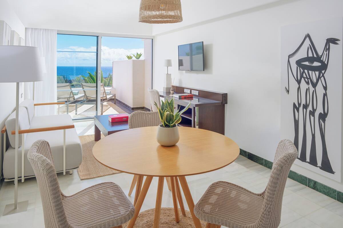 A modern room with a round wooden table, wicker chairs, a potted plant, a flat-screen TV, and a large window offering a view of the ocean.