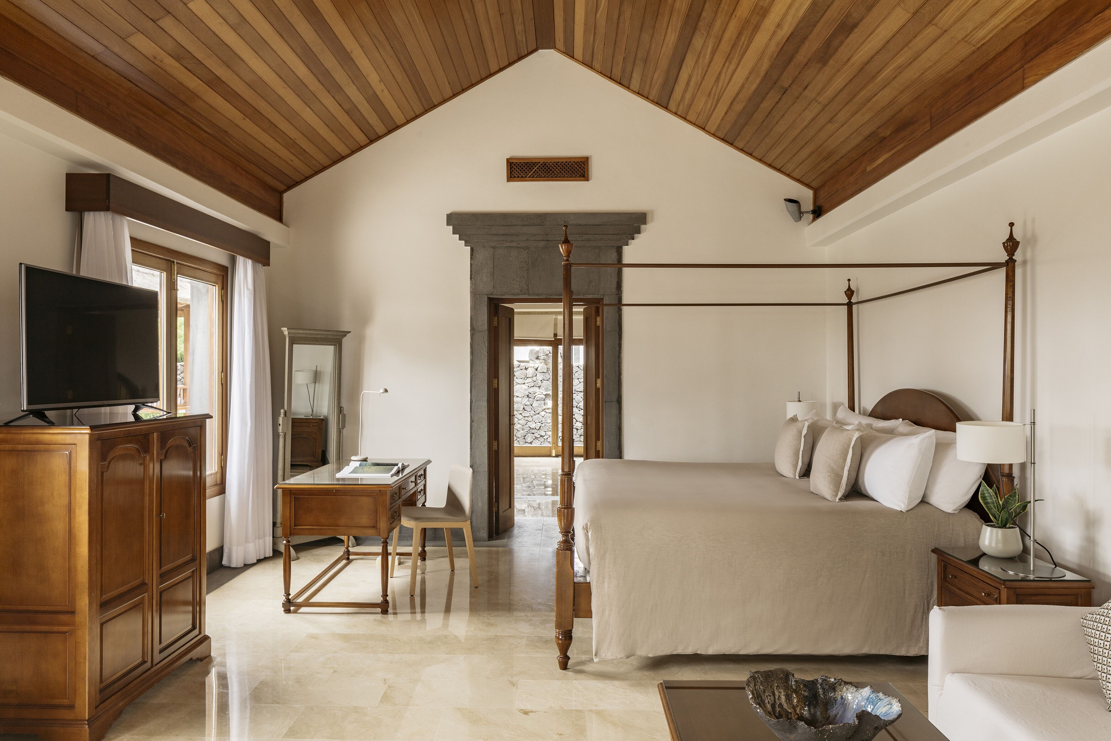 A four-poster bed with a headboard, a television on a wooden cabinet, a desk with a chair, and a stone wall in the background.